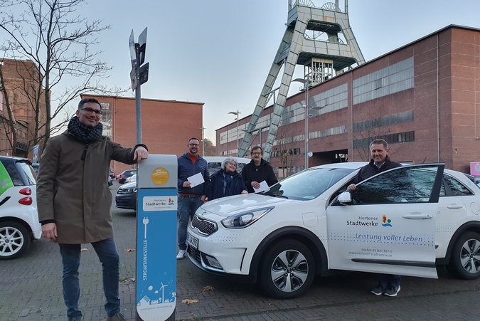 Ein Mann steht an einer Ladestation vor einer Zeche, an der ein weißes Elektroauto geladen wird 