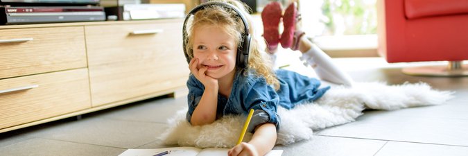 Mädchen liegt fröhlich mit dem Bauch auf dem Boden und malt ein Heft aus. Dabei hört sie Musik und schaut verlegen zur Seite. 