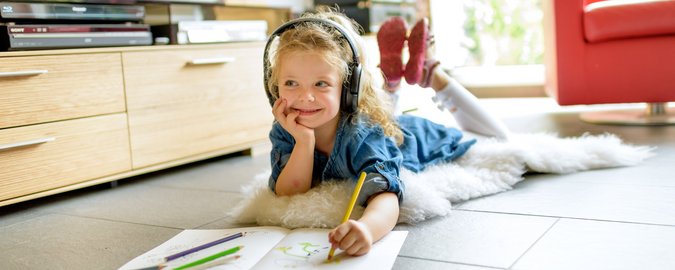 Mädchen liegt fröhlich mit dem Bauch auf dem Boden und malt ein Heft aus. Dabei hört sie Musik und schaut verlegen zur Seite. 