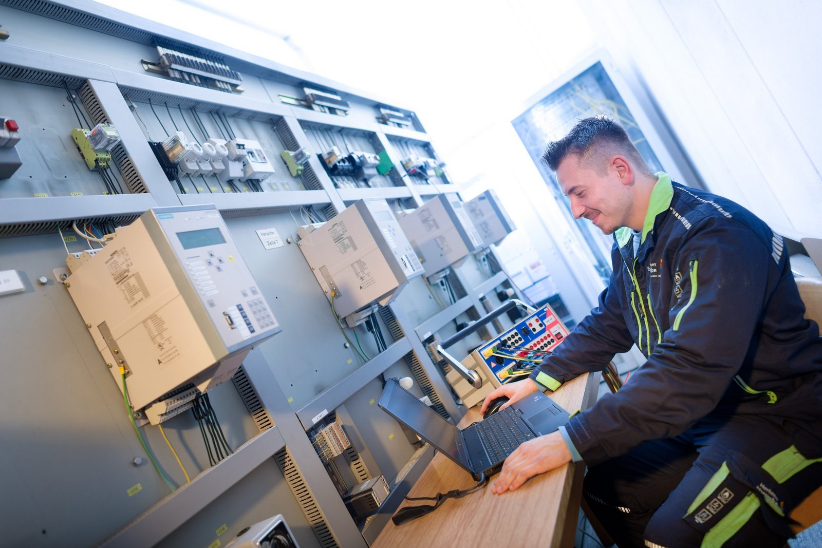 Junger Mann in dunkler Arbeitskleidung in Elektrowerkstatt vor Laptop sitzend