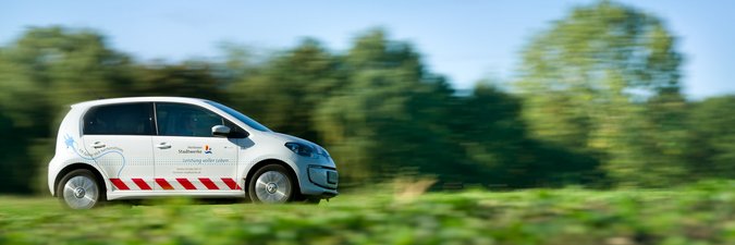 Ein weißes Elektroauto fährt schnell von links nach rechts über eine Landstraße voll grün.