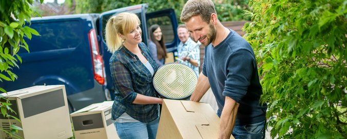 verschiedene Personen, die Kisten eines Umzugs aus einem blauen Bulli Richtung Haus tragen