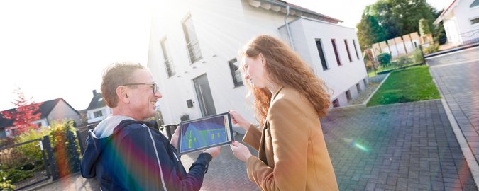 Ein Mann zeigt einer Frau die Gebäudethermografie an seinem iPad. Sie stehen vor einem Haus, das von der Sonne bestrahlt wird.