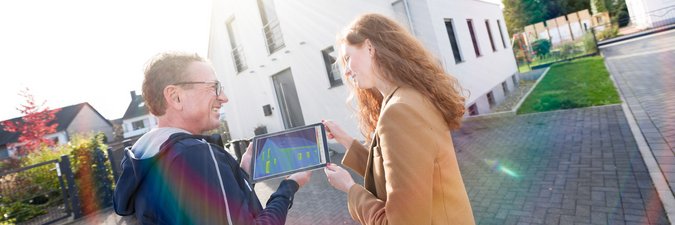 Ein Mann zeigt einer Frau die Gebäudethermografie an seinem iPad. Sie stehen vor einem Haus, das von der Sonne bestrahlt wird.