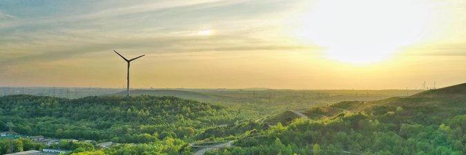 Ein schöner, mit Wäldern, Wiesen und wenigen Straßen bedeckter Berg steht im Vordergrund eines Sonnenuntergang-Panoramas. Links im Hintergrund ein Windrad.