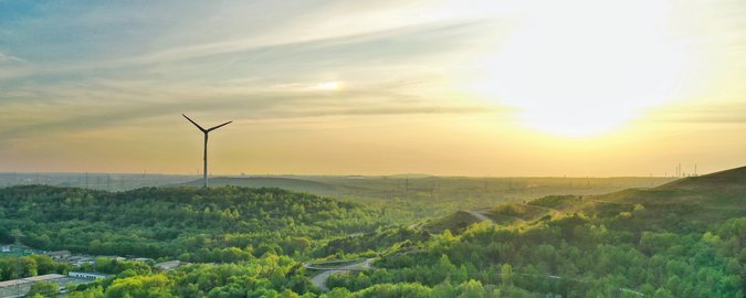 Ein schöner, mit Wäldern, Wiesen und wenigen Straßen bedeckter Berg steht im Vordergrund eines Sonnenuntergang-Panoramas. Links im Hintergrund ein Windrad.