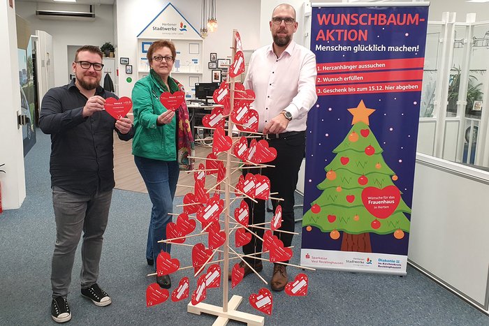 3 Personen stehen vor einem Holzgestell in Form eines Weihnachtsbaumes an dem rote Stiefelsäckchen hängen. Daneben steht ein 2m Plakat in rot und blau mit einem grünem Weihnachtsbaum