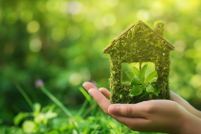 hertenstrom für Wärmepumpen Blätter Hintergrund mit Bokeh Effekt. Hände die ein kleines Holzhaus halten das aus Moos bedeckt ist.