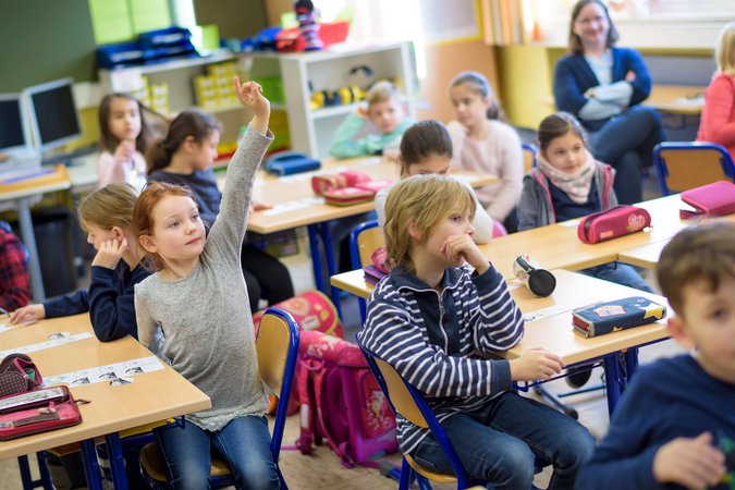 Unterrichtssituation von Grundschülern im Klassenzimmer, ein Kind hebt die Hand hoch und meldet sich