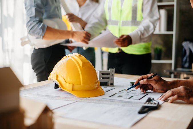 Baustellen Informationen Mehrere Männer sitzen oder stehen um einen Tisch auf dem Baupläne liegen. Im Fokus ist der gelbe Bauarbeiterhelm auf dem Tisch.