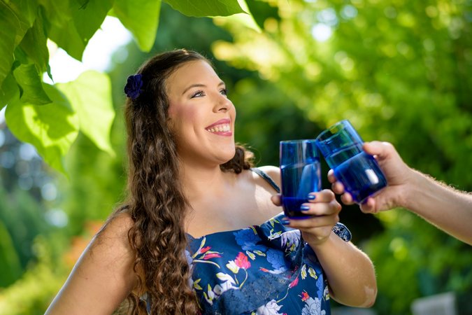 Eine Frau mit braunen Locken steht in einem blauen Sommerkleid mit Blumen darauf unter einem Baum und stößt mit jemandem mit zwei farblich passenden blauen Gläsern an.